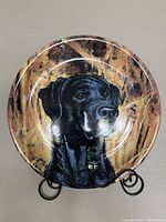 Front view of ceramic plate showing detailed black Labrador retriever portrait against brown marbled background on metal stand.
