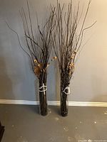 Front view of two tall glass vases filled with brown natural twigs accented with artificial beige flowers and tied with white rope, set against a neutral wall.