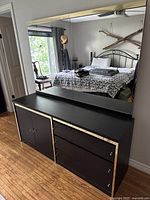 Full view of black wood dresser with large mirror reflecting bedroom. Dresser has three drawers on right, one cabinet door on left with gold trim around edges.