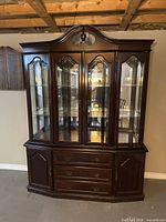 Closed view of the dark brown wood and glass curio cabinet, ornate curved top, three main glass doors with wood framing, three drawers at the bottom center, and two solid wood side cabinets.