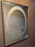 Front view of the wall-mounted decorative mirror showing gold metal frame and etched floral lily design inside an oval border.