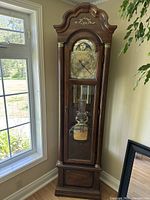 Front view of the tall wooden grandfather clock standing near a window showing its full height and design details.