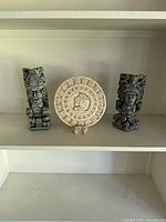 Three Mayan-style clay sculptures displayed on a white shelf: two tribal figure statues and a circular calendar plaque.