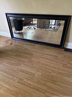 Front angled view of the large rectangular mirror with black wooden frame resting against a wall on a hardwood floor.