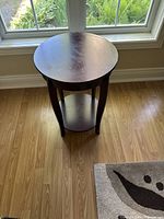Round mahogany-colored MDF side table placed on a hardwood floor near windows, showing the top surface and bottom shelf with natural light reflections.