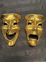 Two brass theatrical style masks lying on a black background, showing comedy (smiling) and tragedy (sad) faces with detailed features and ridged borders.
