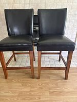 Front view of two black faux leather bar stools with wooden legs on a mixed tile and wood floor.