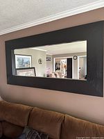 Front view of the rectangular black wood-framed wall mirror mounted above a brown couch reflecting a living room area.