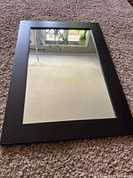 Mirror lying on carpet, showing rectangular shape and black wood frame with clear glass reflecting surroundings.