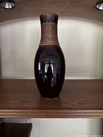 Front view of tall dark brown ceramic vase with twine wrap around the neck placed on wooden shelf.
