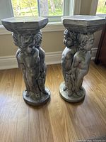 Front and side view of two gray concrete Renaissance style statues depicting cherubic children, each holding a hexagonal top platform, placed on a wooden floor near a window.