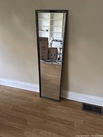 Full view of mirror standing against wall on wooden floor, showing rectangular shape and reflective surface.