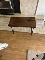 Full top-down view of the wooden table showing the live edge and glossy finish on the plank surface. Positioned on both hardwood and tiled floor areas.