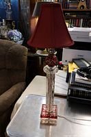 Full view of the entire lamp placed on a table next to books and office equipment, showing the clear glass column base with ruby red spherical accent and red lampshade.