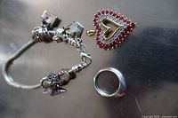 Photo showing all three pieces of jewelry: bracelet with charms, ring, and heart pendant on reflective surface.