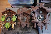Photo showing three vintage wooden cuckoo clocks laid out on a table. Visible clocks feature leaf and bird carvings, various clock faces and missing some parts.