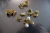 Five pairs of gold coloured clip-on earrings displayed loosely on a flat surface. Shows various designs including lattice, textured hoops and pearl accented earrings.
