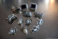 Top view of six pairs of silver colored costume jewelry earrings with various designs including purple stones, faux pearls, and textured metal.
