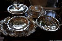 View of 7 silver plated serving pieces on wooden surface including a covered dish, pedestal bowl, flat large platter, and two handled baskets.