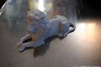 Angled side view of white bisque porcelain lion figurine on reflective surface, showing detail of body and tail