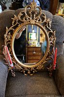 Front view of ornate oval mirror with two attached gold-tone candle sconces and red candles, showing detailed frame and chair background.