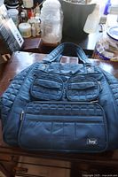 Full front view of blue LUG handbag with quilted sides and multiple zippered pockets, placed on table with other items in background.