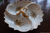 Top angled view showing the white porcelain dish with three divided sections, gold floral designs in each section, scalloped and embossed edges with gold accents, and ornate gold handle in center.