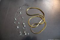 Two costume jewelry necklaces on a reflective surface, one silver with pearls and crystal beads, and one gold flat chain.