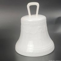 Front and side view of glossy white porcelain bell showing embossed figures and raised inscription near base. Molded loop handle on top visible.