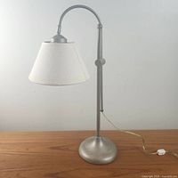 Full view of the adjustable brushed metal desk lamp showing the off-white conical fabric shade, curved adjustable pole, and brushed metal rounded base on a wood surface.