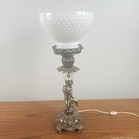 Side view showing ornate brass base with twin cherubs, foliage and beaded detailing; hobnail milk glass shade and crystal element above the base.
