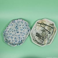 Two vintage metal trays shown front side with decorative prints. One has a blue floral and bird motif, the other a painted street scene with cathedral and trees. Both have shaped, ornate edges.
