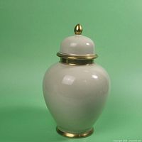 Front view of glossy white porcelain ginger jar with gold-tone accents on lid finial, lid rim, and base against green background.