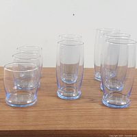 Side view of eight blue-tinted glass tumblers arranged in three groups by height on a wooden surface, showing rounded bases and transparent blue glass.