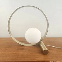 Front view of modern brass ring orb table lamp showing circular brass frame and frosted glass globe shade with wooden surface background.