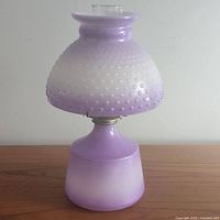 Front view of purple-to-white ombre hobnail oil lamp showing the rounded hobnail patterned glass shade and matching base placed on wooden surface against white wall.