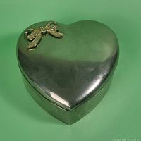Closed heart-shaped silver-plated jewellery box with raised gold-tone bow on lid against green background.