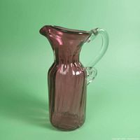 Full side view of a small cranberry glass carafe showing ribbed vertical panel body, flared spout, and clear applied handle.