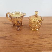 Front view showing creamer with three shell-like feet and scalloped rim beside the lidded sugar bowl with finial top, highlighting amber color and honeycomb textured pattern.