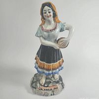 Front view of the ceramic folk woman figurine standing on a stone-like base with the inscription 'CALABRIA FOLK'. Shows detailed painted dress, tambourine and headscarf.