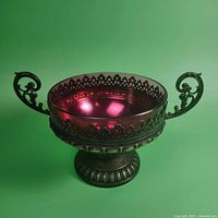 Front angled view of cranberry glass bowl set in ornate cast metal holder with scroll handles and pierced gallery rim, showing overall design and condition.