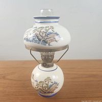 Front angle image of the vintage Japanese ceramic oil lamp showing the rounded ceramic base and upper reservoir with blue accents and scenic hand-painted mountain landscapes.