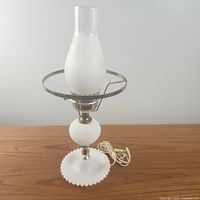 Front angle view showing the full height of the vintage white milk glass hobnail electric lamp with frosted glass chimney, metal support ring, and corded plug lying on the wooden surface.