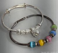 Two sterling silver bracelets shown: one light silver polished with decorative beads and charm, one darker silver bangle with colorful beads and hook clasp
