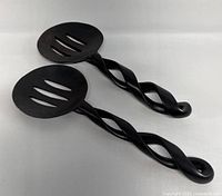 Pair of two hand carved ebony wood serving spoons with twisted handles and slotted heads shown on plain background.