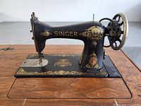 Side view of black Singer sewing machine fixed in wooden table. Machine has gold decals and hand wheel on right side.