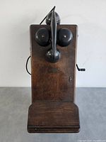Front view of antique wooden wall phone showing two metal bells, mouthpiece, and wooden body.