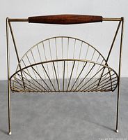 Front view of the mid-century modern style magazine holder stand with brass wire basket and a top teak handle, showing the semi-circular shaped wire rack and four thin legs with white caps.