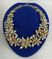 Full view of the floral necklace on a blue display stand showing multiple flowers with rhinestones in various colors.