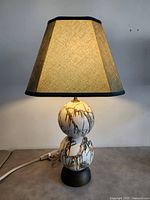 Vintage ceramic accent table lamp with beige tapered fabric shade, double-sphere ceramic base with drip glaze design in brown, green and gold on white, on dark pedestal base, plugged in on a grey surface.